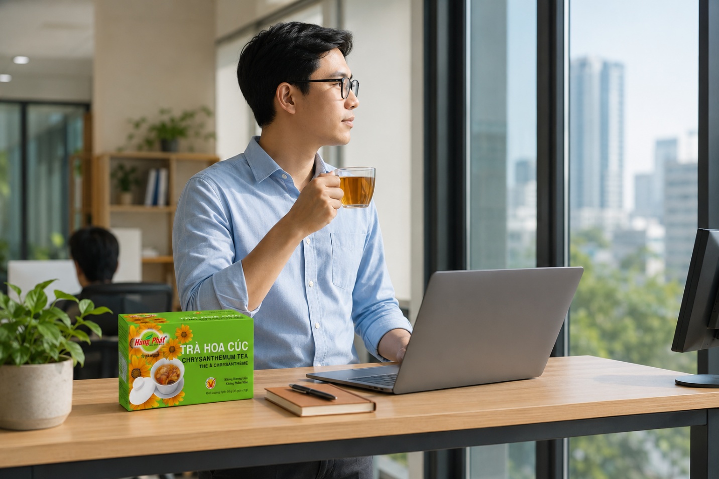 hộp Trà Hoa Cúc Hùng Phát đặt trên góc bàn làm việc, cạnh laptop và tách trà thủy tinh đang bốc khói nhẹ
